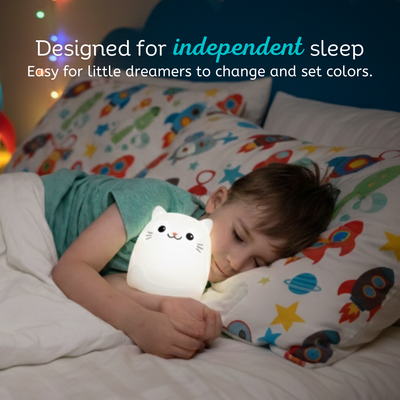 Child lying in bed with a white cat-shaped night light, surrounded by colorful bedding.